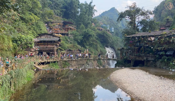 a trail of tourists trekking along a river with bamboo houses in background